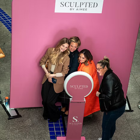 Four women smiling and posing together at a Sculpted by Aimee photo booth with a pink backdrop.