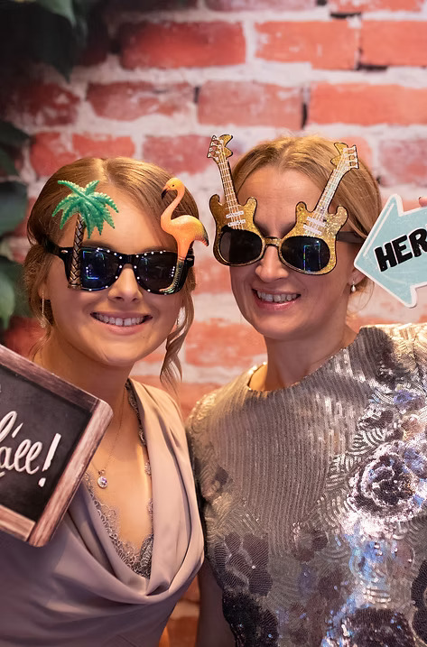 Two women smiling at a photo booth wearing novelty sunglasses shaped like palm trees, flamingos, and guitars, holding playful props against a brick wall backdrop.