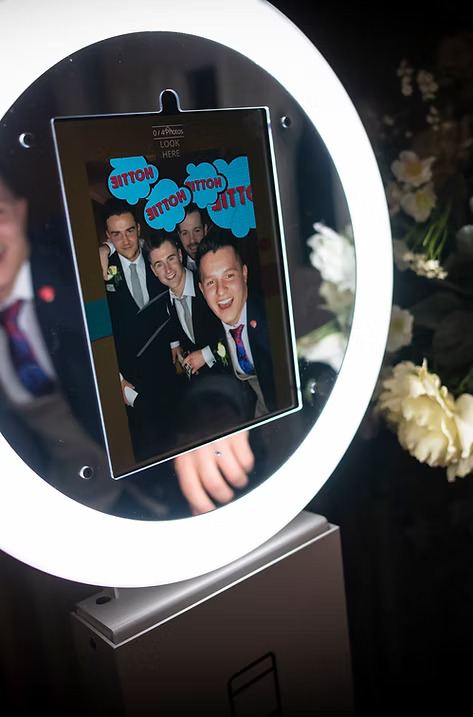 Close-up of a ring light photo booth screen showing a group of men in suits smiling with fun 'Hottie' graphic overlays.