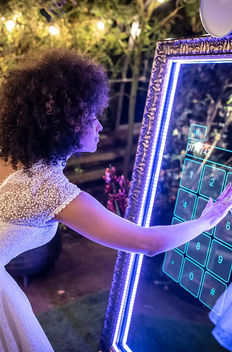 Bride in a white wedding dress interacting with an illuminated mirror photo booth touchscreen during an outdoor evening reception.