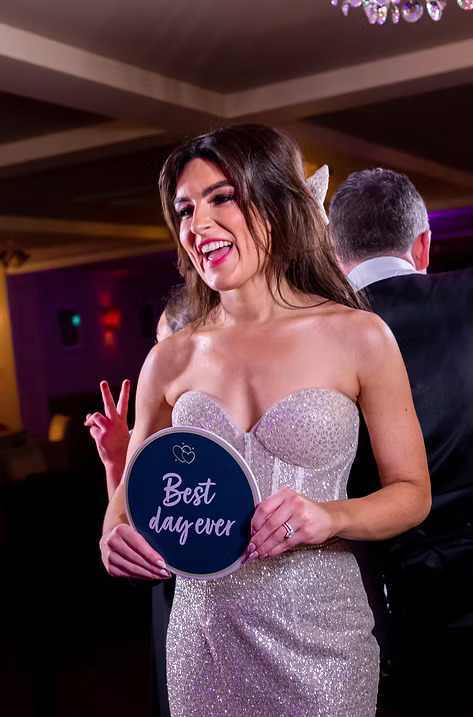 Smiling bride in a sparkling strapless gown holding a round sign that reads 'Best day ever' at her wedding reception.