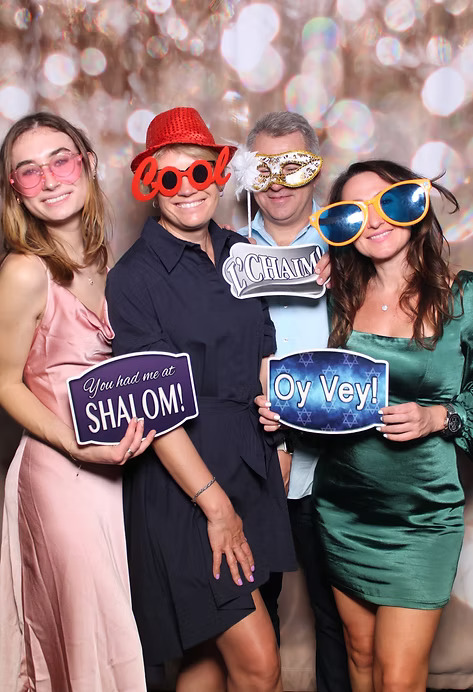 Group of four people posing at a photo booth with oversized sunglasses, hats, and funny signs including 'Shalom,' 'Oy Vey,' and 'L’Chaim.