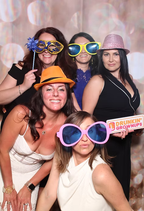 Group of women at a photo booth wearing oversized glasses and colorful hats, holding a sign that says 'Drunkin’ Grownups.