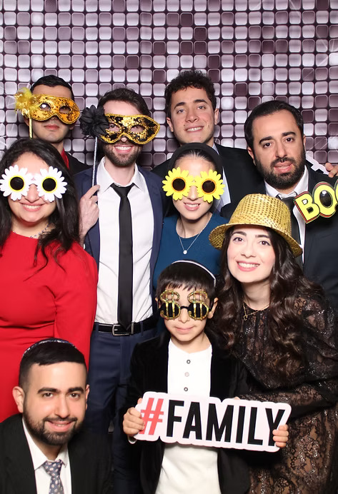 Group of family and friends posing at a photo booth with fun props including oversized glasses, hats, masks, and a #Family sign.