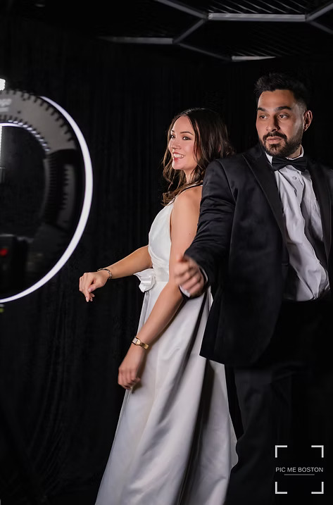 Bride and groom in formal attire posing together in front of a ring light at a photo booth with a black backdrop.