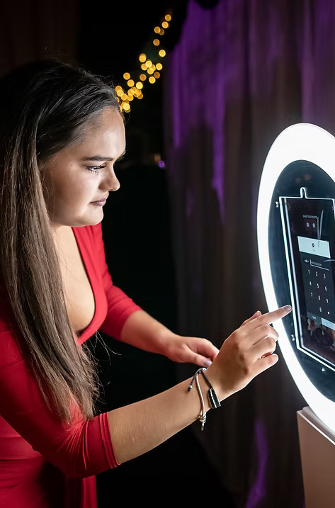 Young woman in a red dress using the touchscreen on a ring light photo booth at an event.