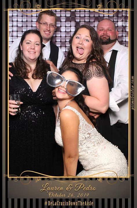 Group of wedding guests, including the bride in a white lace gown, posing at a photo booth with oversized glasses and fun expressions. Frame reads 'Lauren & Pedro, October 26, 2019.