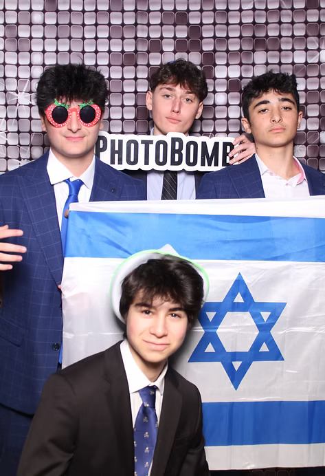 Four young men in suits posing at a photo booth, holding an Israeli flag and a 'Photobomb' sign.