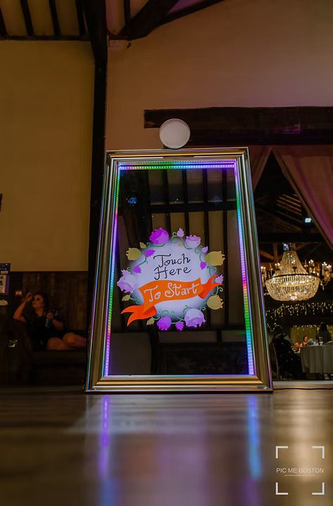 Interactive mirror photo booth with colorful LED lights and a 'Touch Here to Start' welcome screen at an elegant indoor event venue.