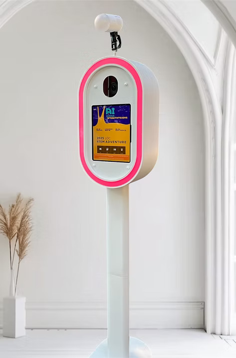 Modern photo booth kiosk with a touchscreen and pink LED border, set up indoors against a white arched wall.