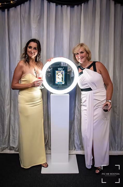 Two women in elegant dresses posing with glasses of wine beside a ring light photo booth at an event.