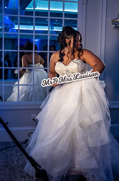 Bride in a sparkling white gown holding a 'Mr & Mrs Louison' sign while posing at her wedding reception.
