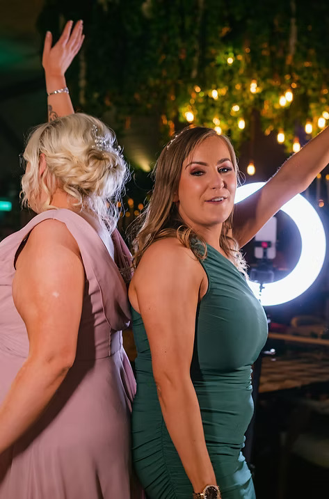 Two women in formal dresses posing back-to-back in front of a ring light at an event photo booth.