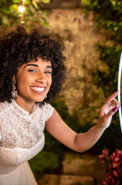 Smiling woman in a white lace dress posing playfully in front of a ring light at an outdoor event.