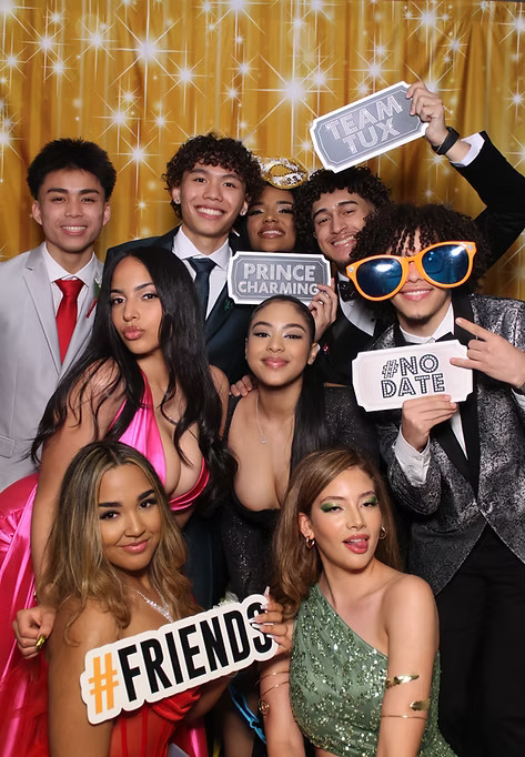 Group of friends dressed in formal prom outfits posing at a photo booth with fun props and signs including '#Friends,' 'Prince Charming,' 'Team Tux,' and '#No Date.