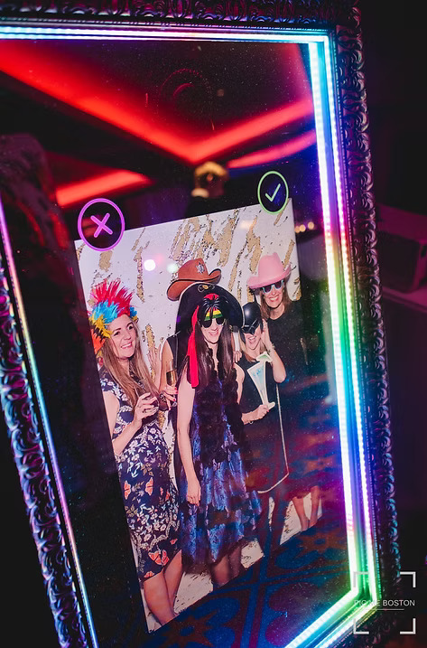 Interactive mirror photo booth showing a group of guests in fun hats and costumes posing for a picture with colorful LED lights around the frame.