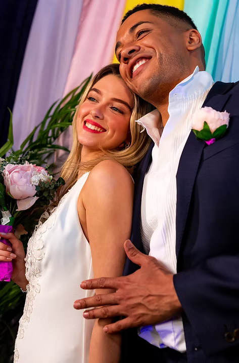 Smiling bride holding a bouquet and leaning on the groom, both dressed elegantly in front of a colorful backdrop.