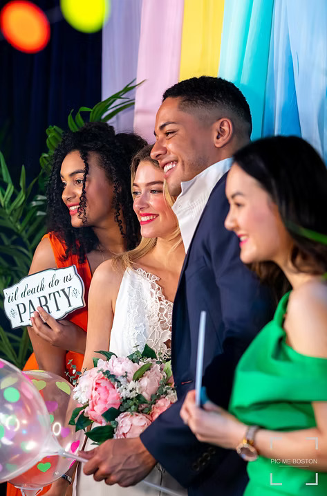 Group of wedding guests posing happily with props and flowers in front of a colorful backdrop at a photo booth.