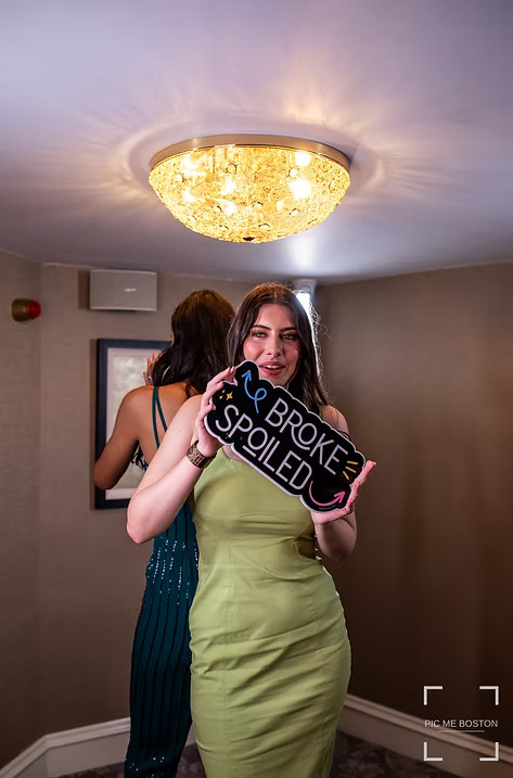 Guest in a green dress posing with a playful 'Broke but Spoiled' photo booth sign at an event.