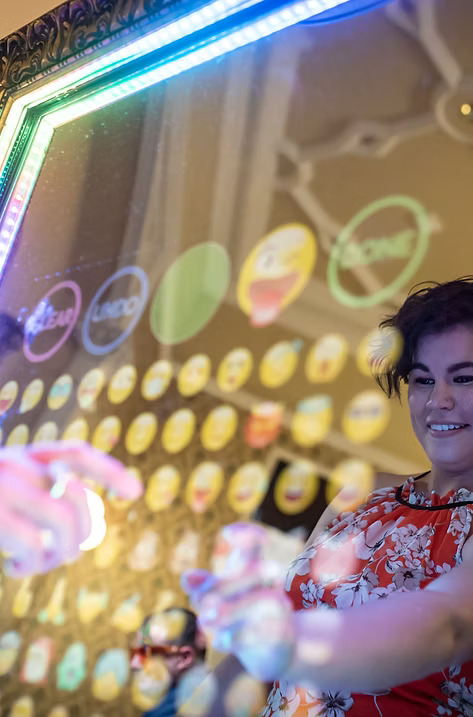 Guest smiling while using a touchscreen mirror photo booth with colorful emoji options and LED lights around the frame.
