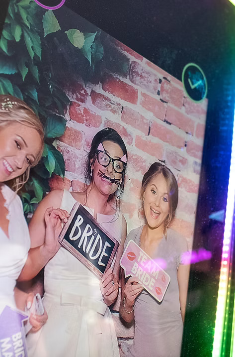 Three women posing in a mirror photo booth, with the bride holding a 'Bride' sign and her friends holding fun props against a brick wall backdrop.