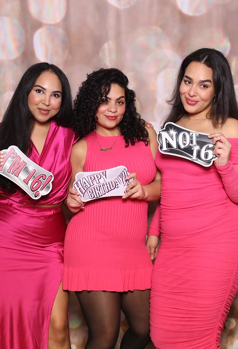 Three young women in pink dresses posing at a photo booth with birthday signs that say 'I’m 16,' 'Happy Birthday,' and 'Not 16.