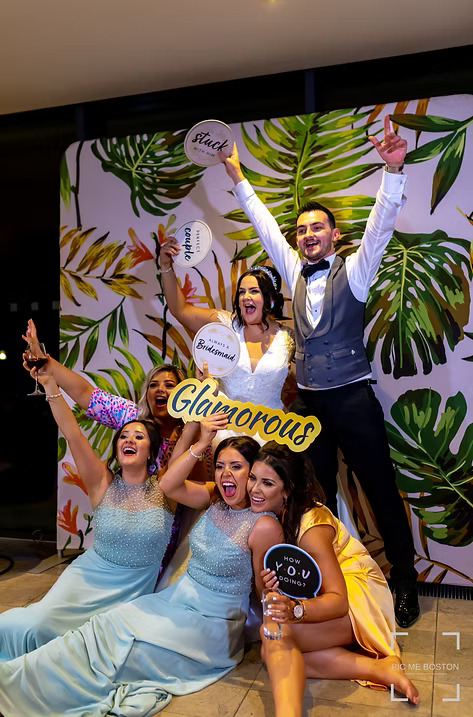 Bride, groom, and friends celebrating in front of a tropical-themed photo booth backdrop, holding fun props with words like 'Glamorous' and 'Bridesmaid'.