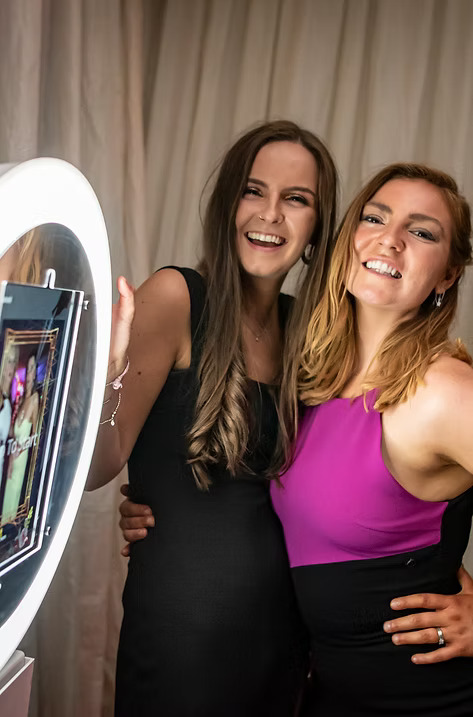 Two women smiling and posing together in front of a ring light photo booth during an event.